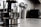 A modern kitchen countertop features a glass electric kettle with a stainless steel base, filled partially with water. Nearby, a white container holds several metallic cooking utensils such as spatulas and ladles. In the background, a microwave oven and other kitchen appliances are slightly blurred.