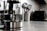 A modern kitchen countertop features a glass electric kettle with a stainless steel base, filled partially with water. Nearby, a white container holds several metallic cooking utensils such as spatulas and ladles. In the background, a microwave oven and other kitchen appliances are slightly blurred.