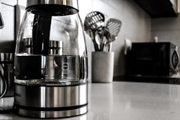 A modern kitchen countertop features a glass electric kettle with a stainless steel base, filled partially with water. Nearby, a white container holds several metallic cooking utensils such as spatulas and ladles. In the background, a microwave oven and other kitchen appliances are slightly blurred.
