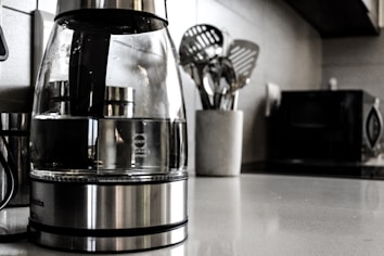 A modern kitchen countertop features a glass electric kettle with a stainless steel base, filled partially with water. Nearby, a white container holds several metallic cooking utensils such as spatulas and ladles. In the background, a microwave oven and other kitchen appliances are slightly blurred.