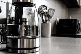 A modern kitchen countertop features a glass electric kettle with a stainless steel base, filled partially with water. Nearby, a white container holds several metallic cooking utensils such as spatulas and ladles. In the background, a microwave oven and other kitchen appliances are slightly blurred.
