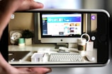A workspace featuring a computer monitor displaying a design interface is seen through a smartphone camera. The desk has a keyboard, mouse, Starbucks cup, clock, and a plant, alongside other small office items. The forward perspective captures the clarity of the workspace setup, implying a focus on technology and design.
