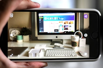 A workspace featuring a computer monitor displaying a design interface is seen through a smartphone camera. The desk has a keyboard, mouse, Starbucks cup, clock, and a plant, alongside other small office items. The forward perspective captures the clarity of the workspace setup, implying a focus on technology and design.