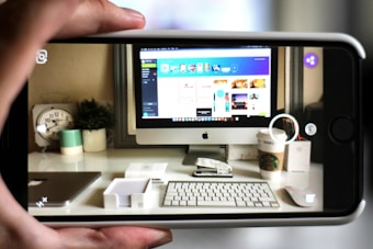 A workspace featuring a computer monitor displaying a design interface is seen through a smartphone camera. The desk has a keyboard, mouse, Starbucks cup, clock, and a plant, alongside other small office items. The forward perspective captures the clarity of the workspace setup, implying a focus on technology and design.