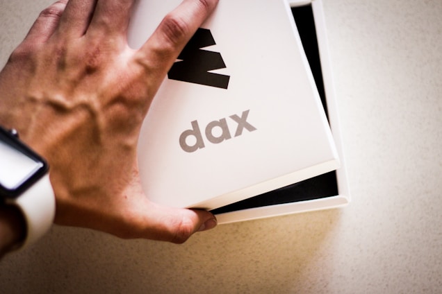 A hand wearing a white watch is opening a white box with the word 'dax' printed on it. The box is partially open, revealing a black interior.