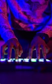 Hands are actively interacting with an electronic music instrument or DJ controller. The scene is illuminated by vibrant neon lighting, casting shades of blue, pink, and purple.