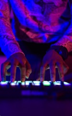Hands are actively interacting with an electronic music instrument or DJ controller. The scene is illuminated by vibrant neon lighting, casting shades of blue, pink, and purple.