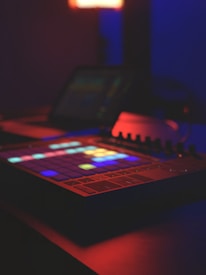 A digital music production device with colorful, illuminated square pads sits in a dimly lit room. The ambient light casts a blue and red glow, enhancing the futuristic and creative atmosphere.