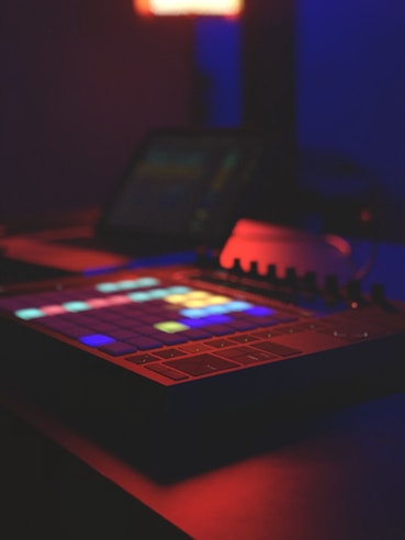 A digital music production device with colorful, illuminated square pads sits in a dimly lit room. The ambient light casts a blue and red glow, enhancing the futuristic and creative atmosphere.