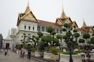 An ornate building with a traditional architectural style featuring golden spires, detailed carvings, and a red and gold roof. The foreground has well-manicured trees and a paved pathway with people walking around, suggesting a popular or tourist area. The scene exudes a sense of historical and cultural significance.