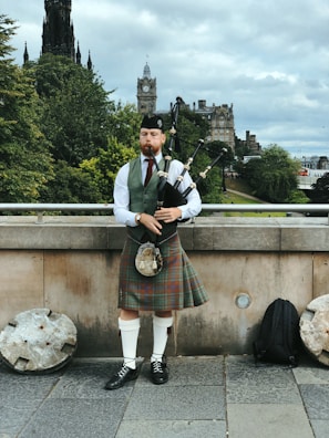 A man in traditional Scottish attire is playing the bagpipes on a city street. He wears a tartan kilt, white shirt, vest, and a hat. Behind him, historic buildings and lush green trees are visible, along with a stone structure that resembles a clock tower.