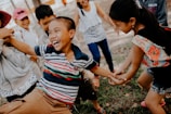 A group of children are playing and laughing outdoors. One child, wearing a striped shirt, is being playfully pulled by other children. The scene is animated and full of energy, with everyone appearing happy and engaged in the activity.