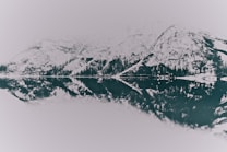A serene mountain landscape with snow-covered peaks reflected on the surface of a still, clear lake. The scene is in monochrome, enhancing the contrast and reflection of the rugged mountains and evergreen trees lining the water's edge. A misty ambiance envelops the background, creating a sense of tranquility.