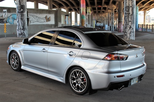 A silver Mitsubishi Lancer Evolution is parked in an urban environment under an overpass, surrounded by concrete pillars adorned with street art and graffiti. The car's sleek design features sporty rims and dark tinted windows.