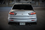 A silver sports car is prominently displayed from the rear, with distinctive tail lights and a dual exhaust. The car is parked in a dimly lit area, possibly a garage or an underpass, with murals visible in the background. The license plate indicates the vehicle is registered in Colorado.