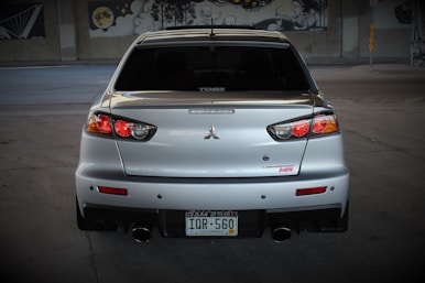 A silver sports car is prominently displayed from the rear, with distinctive tail lights and a dual exhaust. The car is parked in a dimly lit area, possibly a garage or an underpass, with murals visible in the background. The license plate indicates the vehicle is registered in Colorado.