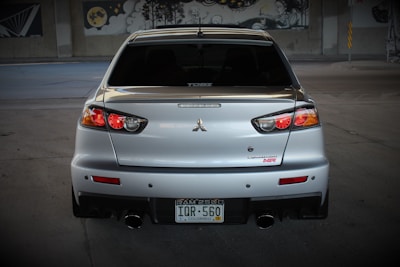 A silver sports car is prominently displayed from the rear, with distinctive tail lights and a dual exhaust. The car is parked in a dimly lit area, possibly a garage or an underpass, with murals visible in the background. The license plate indicates the vehicle is registered in Colorado.