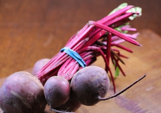 A bunch of fresh purple beets with long, vibrant red stems, secured together with a blue elastic band, rests on a wooden surface.