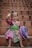 A young child is sitting on brick steps, wearing vibrant traditional clothing. The outfit features intricate patterns in bold colors like pink, green, and purple. The child is smiling and waving, holding a decorative hat with similar colorful designs.