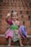 A young child is sitting on brick steps, wearing vibrant traditional clothing. The outfit features intricate patterns in bold colors like pink, green, and purple. The child is smiling and waving, holding a decorative hat with similar colorful designs.
