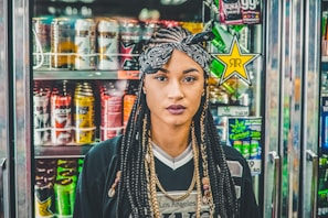 A woman with braided hair and wearing a headband stands in front of a beverage cooler filled with colorful energy drink cans. The background features various brands with vibrant and eye-catching packaging designs.