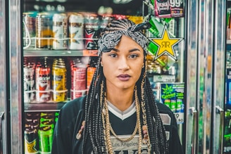 A woman with braided hair and wearing a headband stands in front of a beverage cooler filled with colorful energy drink cans. The background features various brands with vibrant and eye-catching packaging designs.