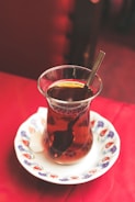 A glass of black tea sits on an ornately decorated white saucer, with a small cube of sugar beside it. The red tablecloth provides a vibrant backdrop, complementing the dark amber color of the tea. A shiny metal spoon rests inside the glass, reflecting light from the surroundings.