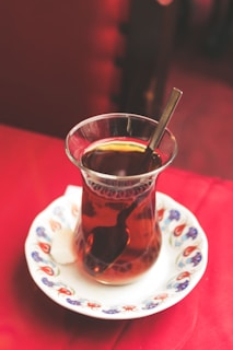 A glass of black tea sits on an ornately decorated white saucer, with a small cube of sugar beside it. The red tablecloth provides a vibrant backdrop, complementing the dark amber color of the tea. A shiny metal spoon rests inside the glass, reflecting light from the surroundings.