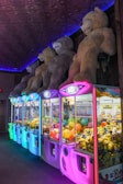 A row of neon-lit claw machines featuring various prizes, including colorful balls and toy minions. Above the machines, large plush teddy bears are displayed, casting a playful and inviting atmosphere.