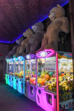 A row of neon-lit claw machines featuring various prizes, including colorful balls and toy minions. Above the machines, large plush teddy bears are displayed, casting a playful and inviting atmosphere.