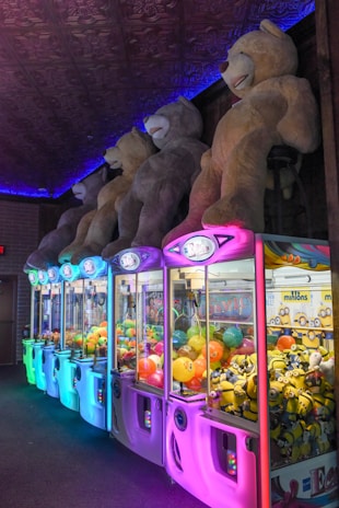 A row of neon-lit claw machines featuring various prizes, including colorful balls and toy minions. Above the machines, large plush teddy bears are displayed, casting a playful and inviting atmosphere.