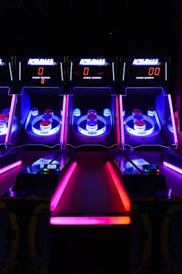 A row of brightly lit skee-ball machines with neon lights in a dark arcade setting. Each machine has a digital score display, currently showing scores of zero. The playing area of each machine is illuminated with purple and blue lighting, highlighting the targets and scoring rings.
