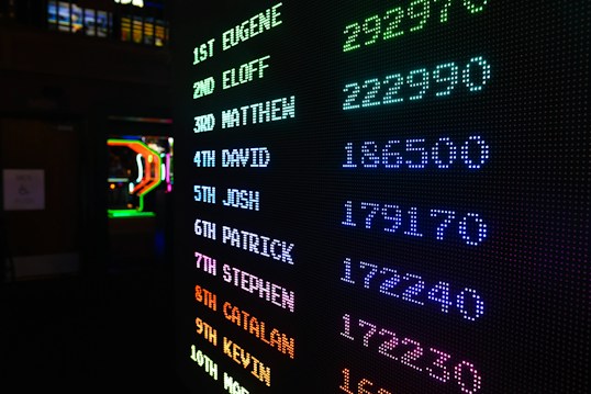 A digital leaderboard displaying names and scores in vibrant colors like green, blue, purple, and orange. The display is in a dark setting, possibly an arcade, and features a bright LED design.