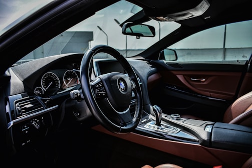 The interior of a luxury car with a focus on the steering wheel and dashboard. The dashboard features a speedometer and other gauges, all of which are detailed and sleek. The gear shift and other controls are positioned on a polished center console. The seats and interior lining are made of rich brown leather, adding to the sophisticated ambiance.