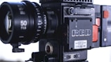 A professional digital cinema camera with a large lens prominently visible. The body features various controls and branding, including a label that reads 'RED'. The design is robust and industrial, with visible screws and a textured surface.