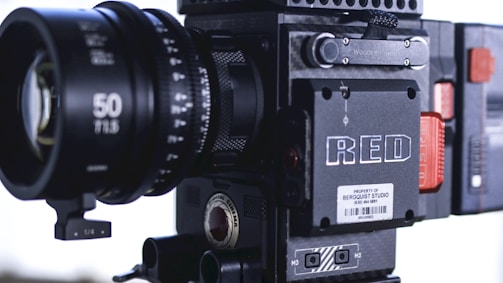 A professional digital cinema camera with a large lens prominently visible. The body features various controls and branding, including a label that reads 'RED'. The design is robust and industrial, with visible screws and a textured surface.
