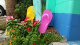 A vibrant display of colorful flip-flops arranged among lush, flowering plants. The backdrop features a teal and blue painted wall. Bright pink, yellow, and aqua flip-flops contrast with the green leaves and multicolored flowers, creating a cheerful and lively scene.