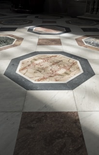 A geometric pattern of octagonal tiles made of marble with various colors such as white, gray, brown, and red. Sunlight is partially illuminating the tiles, creating a contrast between the lit and shadowed areas.