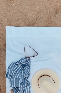 A beach scene with a blue and white striped shirt, a straw hat, and sunglasses on a light blue towel laid out on the sand.