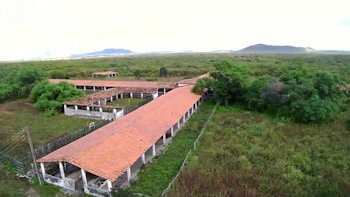 An aerial view of a rural landscape with a series of long, narrow buildings with red-tiled roofs surrounded by lush greenery and trees. The area is expansive, with a fence enclosing the structures. In the background, distant hills or mountains are visible against a bright sky.