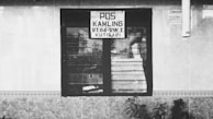 A black and white photograph of a window with a sign above it. The sign reads 'POS KAMLING RT.04-RW.1 KUTISARI.' The window reflects some foliage outside. The building has tiled walls, and there are some plants in front.