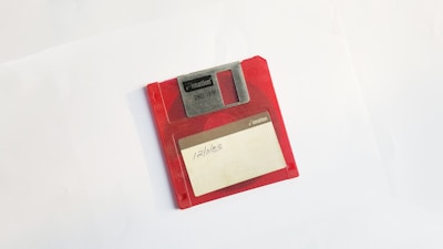 A red floppy disk with a label that reads '12/11/03'. The disk is positioned at a slight angle on a plain white background. It has a visible metal sliding cover and branding that indicates it is a 2HD IBM disk from Imation.