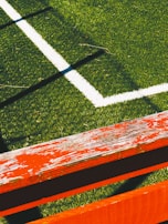 The image captures a section of a sports field with lush green artificial turf and a white boundary line intersecting at a right angle. A weathered wooden bench painted in bright orange with visible peeling paint is positioned diagonally across the foreground, casting shadows on the grass.