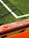 The image captures a section of a sports field with lush green artificial turf and a white boundary line intersecting at a right angle. A weathered wooden bench painted in bright orange with visible peeling paint is positioned diagonally across the foreground, casting shadows on the grass.