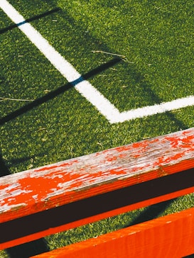 The image captures a section of a sports field with lush green artificial turf and a white boundary line intersecting at a right angle. A weathered wooden bench painted in bright orange with visible peeling paint is positioned diagonally across the foreground, casting shadows on the grass.