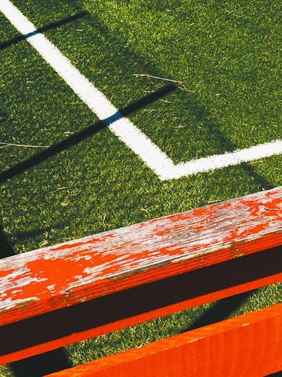 The image captures a section of a sports field with lush green artificial turf and a white boundary line intersecting at a right angle. A weathered wooden bench painted in bright orange with visible peeling paint is positioned diagonally across the foreground, casting shadows on the grass.