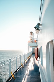 A peaceful scene featuring a ship's deck bathed in warm sunlight with the ocean visible to the left. The deck is lined with white railings and has yellow safety posts, creating long shadows on the deck floor. A red emergency box is mounted on the ship's side, and large ship speakers are attached above it.
