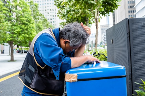 A person wearing a blue uniform is leaning on a blue container with one hand on their head, appearing tired or stressed. The scene is set outdoors in an urban area with green trees and tall buildings in the background.