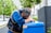 A person wearing a blue uniform is leaning on a blue container with one hand on their head, appearing tired or stressed. The scene is set outdoors in an urban area with green trees and tall buildings in the background.
