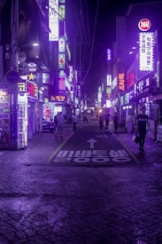 A vibrant urban street scene at night, illuminated by numerous neon signs with Korean characters. The street is bustling with people walking amidst a backdrop of colorful lights and signs. The ground has visible textures and is marked by a painted arrow and Korean text.